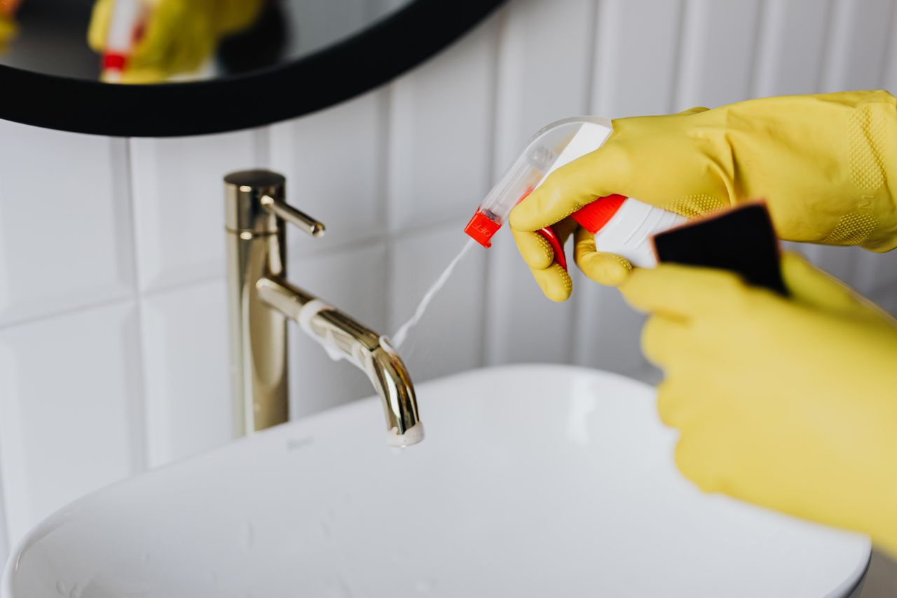 From an overhead view, an anonymous housekeeper in yellow rubber gloves sprays cleaner onto a chrome bathroom faucet.