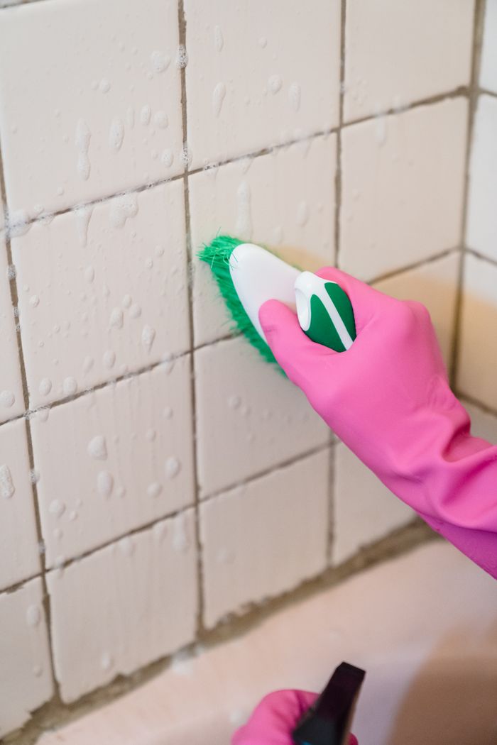 Une main gantée de rose nettoie des carreaux de salle de bain à l'aide d'une brosse verte pour enlever la saleté.